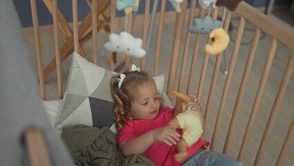 Fototapeta premium Little girl holding a doll and reaching toward a cloud and moon mobile while sitting in wooden crib in a nursery building with geometric pillow and blanket; playful comfort.