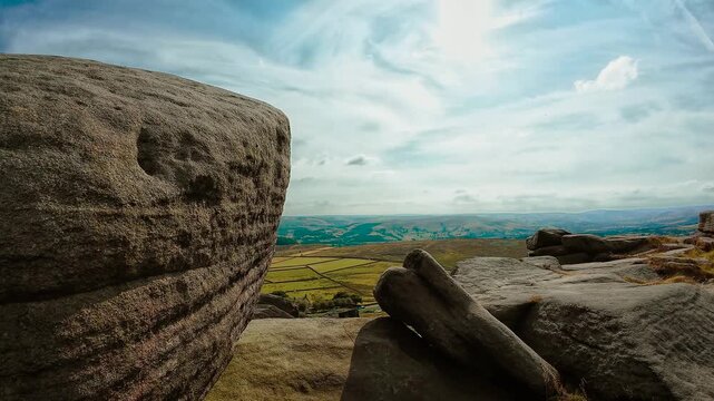 Close granite boulder beside panoramic rural valley under bright cloudscape slide shot. Massive weathered stone with patchwork fields and rolling hills. Rugged rock formations above open countryside