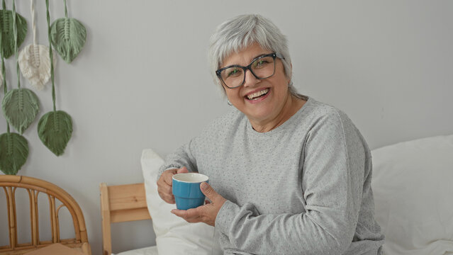 Woman holding blue cup while seated on bed in building, smiling at camera with short gray hair and glasses; calm contentment.