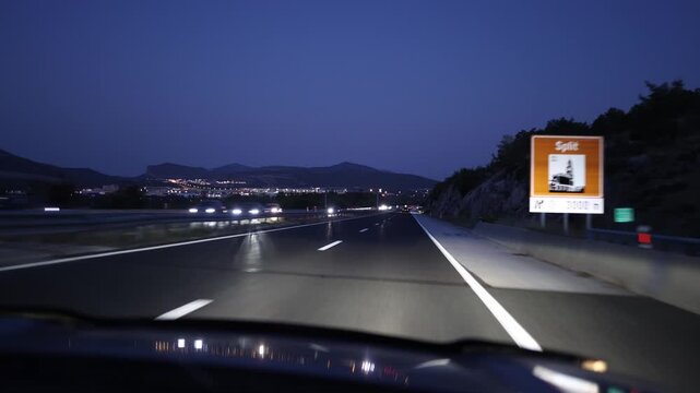 Night highway driving through mountain landscape captures an open motorway illuminated by headlights with distant city lights glowing under a deep blue evening sky beyond