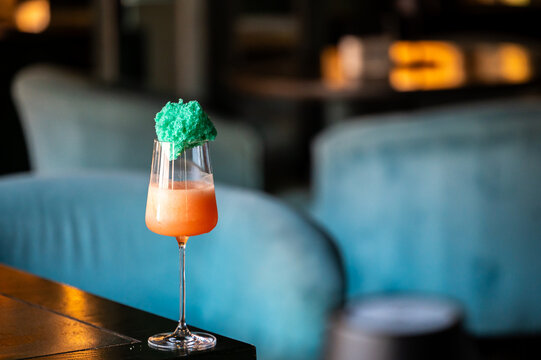 A vibrant cocktail with a pinkish-orange gradient sits on a bar table, topped with a green sponge-like garnish, set against a softly blurred restaurant background with blue velvet seating.