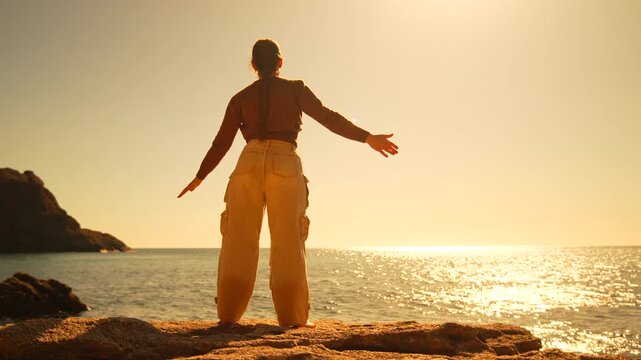 Woman sunset exercise performs light stretching and jumping on a rocky cliff overlooking the ocean for health and mindfulness in nature