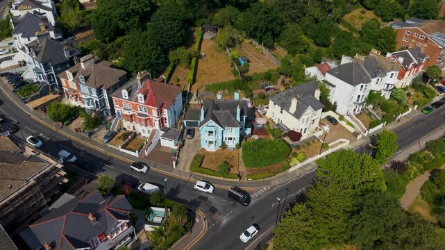 Left aerial pan sweeps over a row of colourful Victorian villas at a road junction beside Alexandra Park in Hastings, with cars passing below and mature gardens bordering the tree-lined park edge.