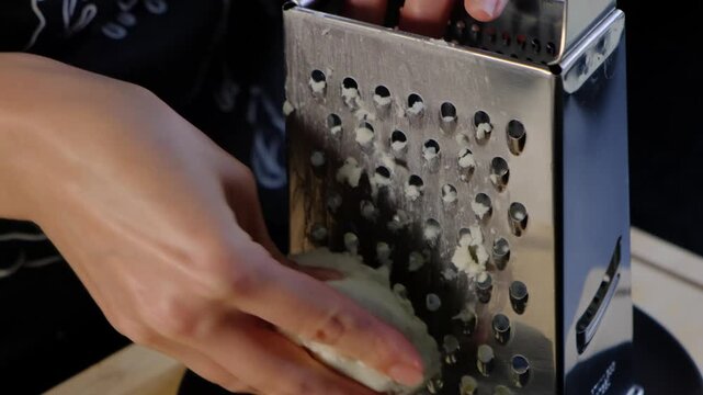 Close-up of hands grating soft cheese or dough through a metal grater plate on an electric appliance in a cooking kitchen, with grated pieces falling into a tray.