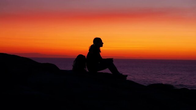 Solitary hiker silhouette seated on rocky coastal bluff with backpack beside figure, facing ocean horizon at vibrant sunset with calm sky, reflective mood, wide scenic view