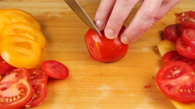 A chef uses a sharp knife to slice ripe red tomatoes on a wooden board near a neat pile of yellow tomato rings, creating precise layers for a healthy organic salad.