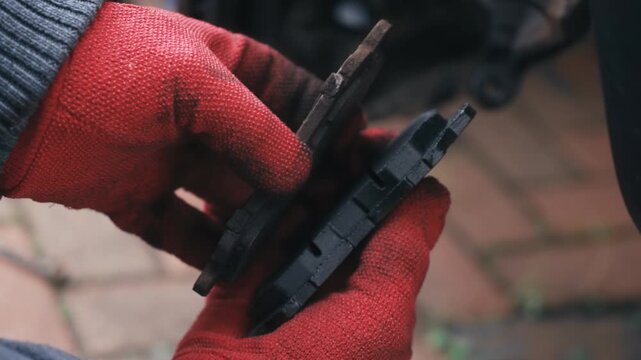 Comparing worn brake pad with new replacement component in gloved hands. Evaluating friction material thickness for vehicle braking maintenance outdoors. Automotive repair inspection focusing on parts
