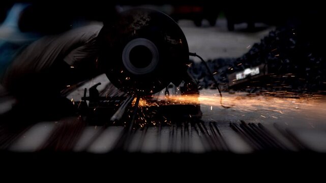 A metalworker cutting steel rebar with an angle grinder while sparks fly in a dark industrial workshop
