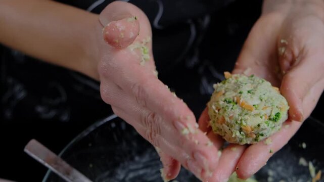 Close-up of hands shaping greenish-white vegetable mixture with orange shreds into patties on dark surface in Cooking kitchen, using metal utensils.