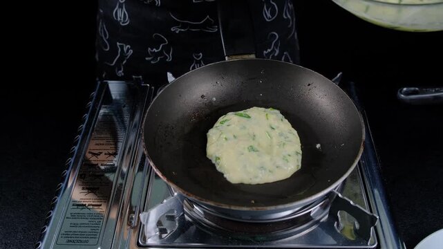 Adult hands in dark patterned clothing stir a pale yellow herb-speckled mixture with a metal spatula in a dark frying pan on a black stovetop, in a kitchen at Cooking location, daytime.