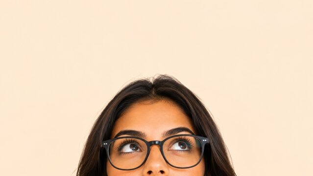 Meio rosto de jovem mulher brasileira usando &oacute;culos de grau olhando para cima. Conceito de exame de vista, oftalmologia e amplo espa&ccedil;o para texto no fundo bege
