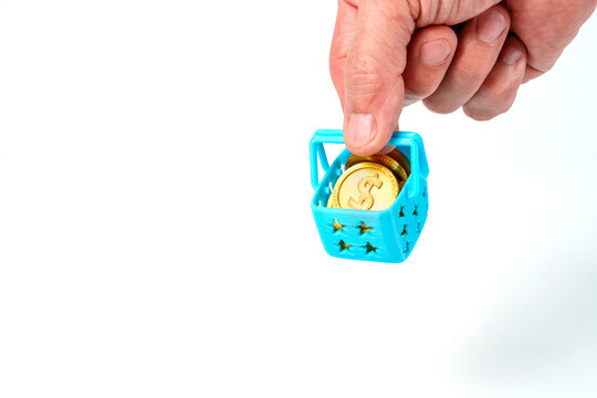 Hand holding a miniature blue shopping basket filled with gold dollar coins on white background. Professional close up representing inflation, consumer spending, retail costs and personal finance.
