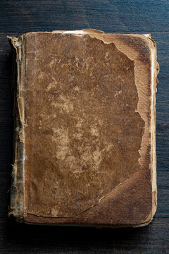 Vintage worn book cover on dark wood, closeup. Top view of an antique book with heavily damaged, weathered brown leather cover lying on dark wooden background