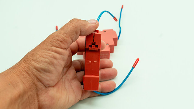 In a workshop, a man holds a red electrical component with two blue wires, a portable fire extinguisher in an electrical panel, and is isolated on a white background. His hands demonstrate careful han