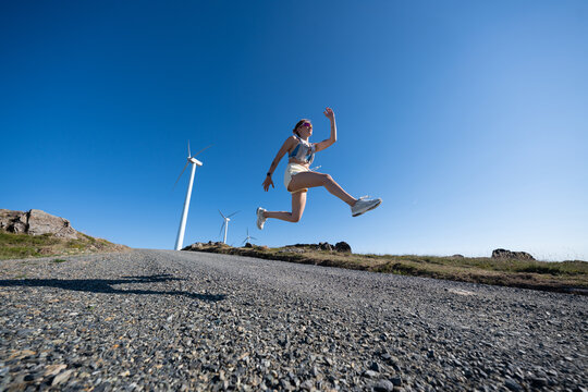 Young white woman jumps outdoors near wind turbines in Galicia, Spain
