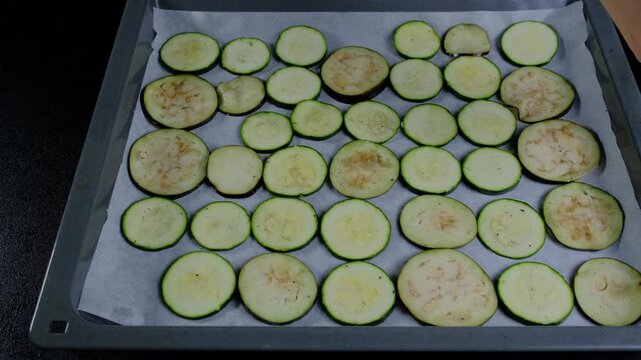 Hand arranging sliced green zucchini on parchment-lined baking tray during food prep in Cooking location, overhead view, daytime kitchen scene.