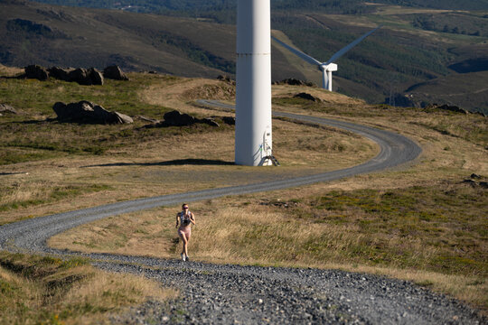A white woman runs on a winding trail by wind turbines in Galicia