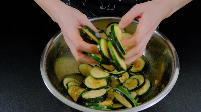 Hands mix sliced zucchini in a stainless steel bowl with marinade during cooking in a kitchen, overhead view.