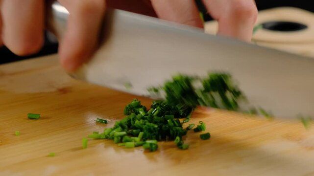 Hands finely mincing green chives with a large knife on a wooden board in a Cooking kitchen, close-up shot during daytime.
