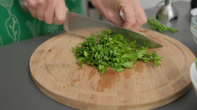 Close-up of hands chopping green herbs on wooden board with chef's knife in Cooking kitchen, bright daylight, food preparation scene.
