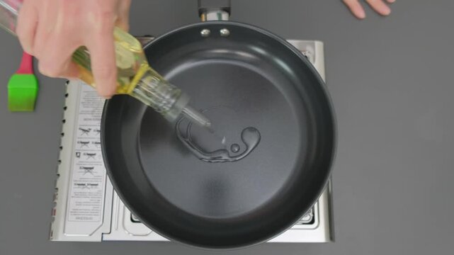Hands pour yellow cooking oil from bottle into dark gray frying pan on gas stove in Cooking kitchen, overhead view, cooking process.