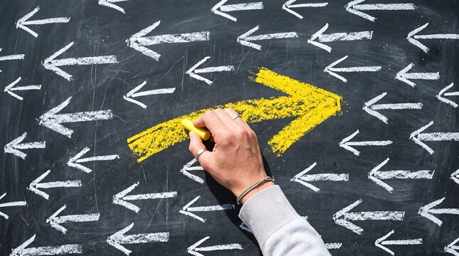 Hand drawing a yellow arrow on a black chalkboard with many white arrows pointing in the opposite direction concept of individuality, leadership, and standing out