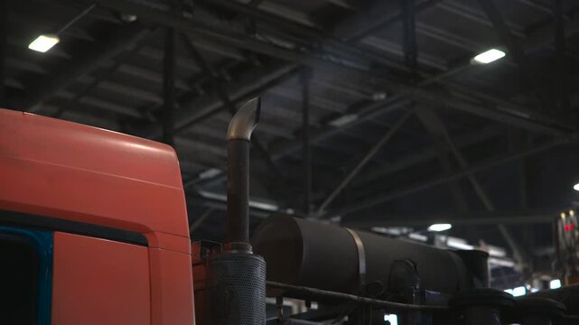 Low angle close view of red truck cab beside vertical exhaust stack inside industrial garage, with faint bluish plume rising near roof trusses and overhead lights