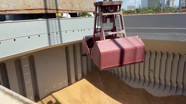 Industrial crane clamshell grab working in ship cargo hold discharging agricultural grain, heavy equipment port logistics operations