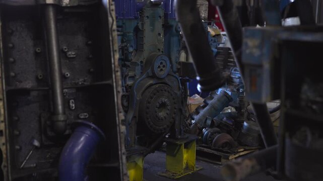 Close-up disassembled blue engine block with exposed flywheel and yellow metal stands inside dim industrial workshop, showing heavy mechanical parts, grime, rust, and service details