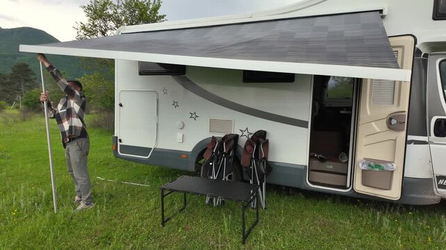 Young man assembling a retractable awning to the Marquis on his motorhome, preparing camp for outdoor recreation in a beautiful green meadow with mountains in the background in summer