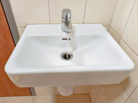 Water flowing from chrome faucet into white ceramic sink drain. Modern bathroom plumbing fixture with running tap water. Concept of hygiene, water conservation and plumbing maintenance