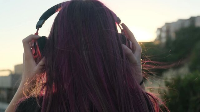 Back view, teenage girl with purple hair puts on red wireless headphones before watching the city sunset. Listening to music, urban sunset, peaceful moments and enjoying the city landscape.