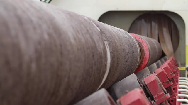 A large steel pipe rotates on powered red roller supports as rough unfinished metal texture and marked joint sections move through bright factory machinery