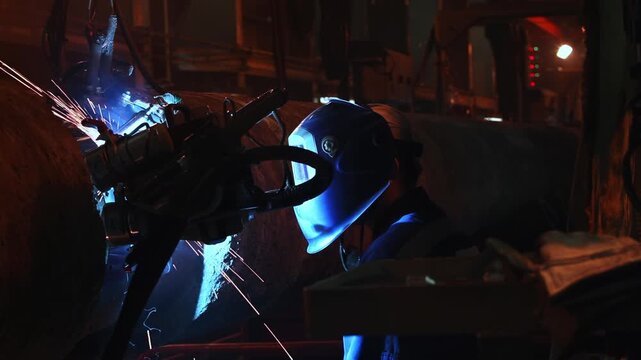 A welder wearing a protective face shield monitors a mechanized welding head joining a large steel pipe as intense blue arc light fills the dark industrial workshop