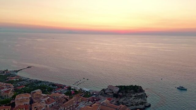 A wide aerial shot capturing the historic town of Tropea at sunset, featuring the Tyrrhenian Sea, coastal cliffs, and the Santa Maria dell Isola monastery at dusk.