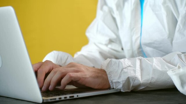 Male scientist in a white hazmat suit is typing on a laptop keyboard, working on research in a laboratory or medical facility with a yellow background, focusing on data entry and analysis