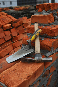 Bricklaying tools including a trowel and mason's hammer resting on a freshly built red brick wall with cement mortar at a construction site.