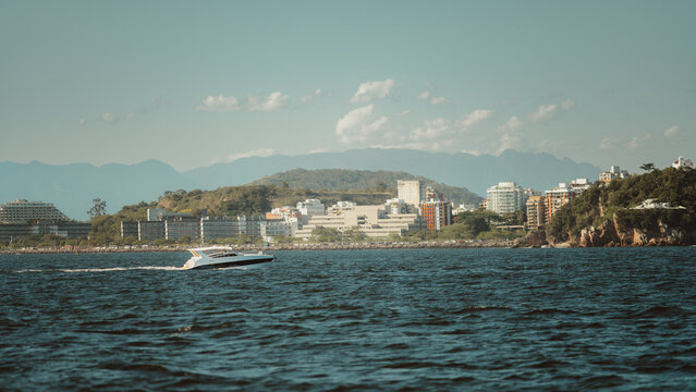 Telephoto view of a sleek motorboat cruising across choppy blue water for a relaxing leisure trip, with a modern urban shoreline and layered mountains rising in the distance