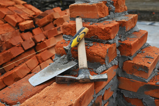 Bricklaying tools including a trowel and mason's hammer resting on a freshly built red brick wall with cement mortar at a construction site. Horizontal photo.