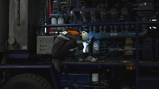 Technician performing focused maintenance on large mobile engine inside vehicle compartment under dim workshop lighting, inspecting lower section, hoses, valves, metal parts, safety gear