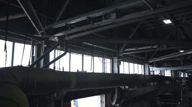 Panoramic industrial roof interior with oversized insulated ventilation duct beneath exposed steel trusses, diagonal support braces, cable trays, weathered metal surfaces, and dim natural light