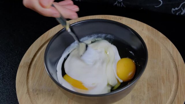 Close-up of hand stirring yellow egg yolks and whites with silver fork in dark gray bowl on wooden board during cooking.