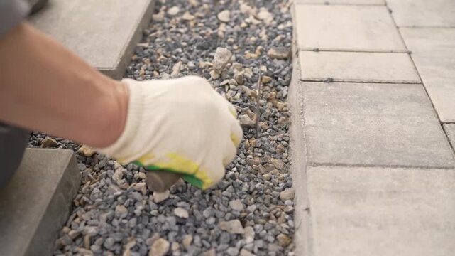 A close-up of a gloved hand laying paving slabs on a gravel base. Neutral gray tones with orange accents for safety. Landscape design, artificial turf.