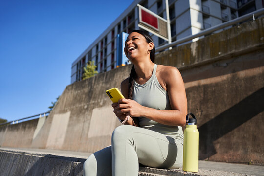 Happy Black Latina athlete sitting outdoors and laughing while checking her smartphone during a workout break, with a reusable water bottle beside her in bright sunlight.