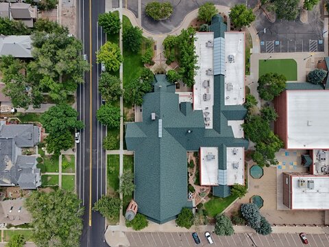 Aerial view of a building with a dark green roof surrounded by lush trees, parking lots, and adjacent streets in Longmont, Colorado, United States.