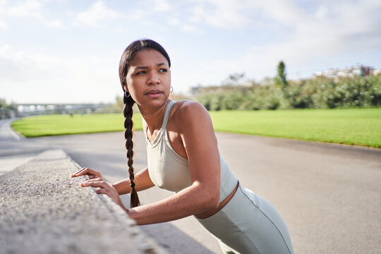 Focused Black Latina athlete leaning against a concrete wall while stretching and looking away in an urban park. Outdoor fitness scene conveying preparation, recovery and concentration.