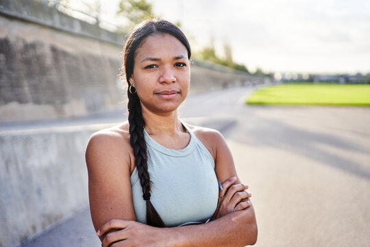 Arms crossed African millennial woman standing outdoors in park after exercise, wearing sportswear and looking confident at camera. Natural light portrait shows strength, balance and active lifestyle.
