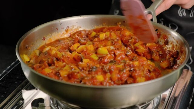 Chef stirring chunky vegetable sauce in large stainless steel pot on stovetop during cooking in kitchen, Cooking location, daytime meal preparation.