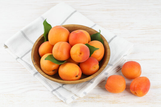 Top view of ripe apricots with green leaves arranged in wooden bowl on on white tile background. Fresh seasonal harvest, summer fruit, healthy eating, dessert, food ingredient