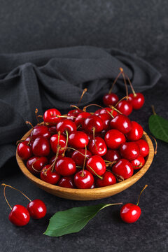 Fresh ripe red sweet cherries in wooden bowl on dark textured stone background with green leaves and black linen towel. Summer fruit, berry, harvest, healthy organic food